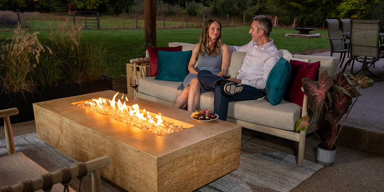 An outdoor patio space with a FlameCraft Tuscan gas fire pit and a couple sitting nearby on an outdoor sofa