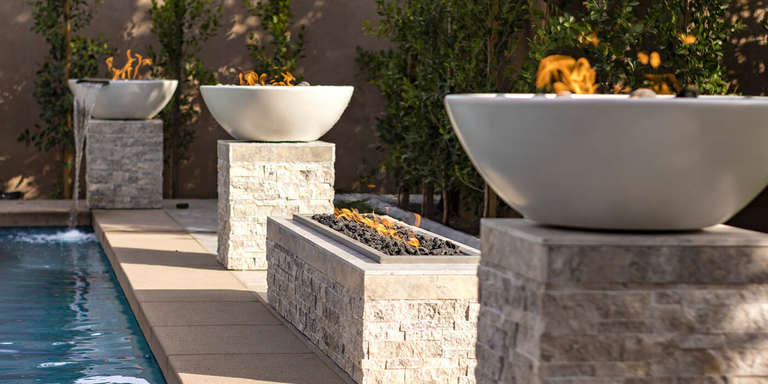 Three gas fire bowls and a linear gas fire pit placed near the edge of an in-ground pool in a luxury modern backyard