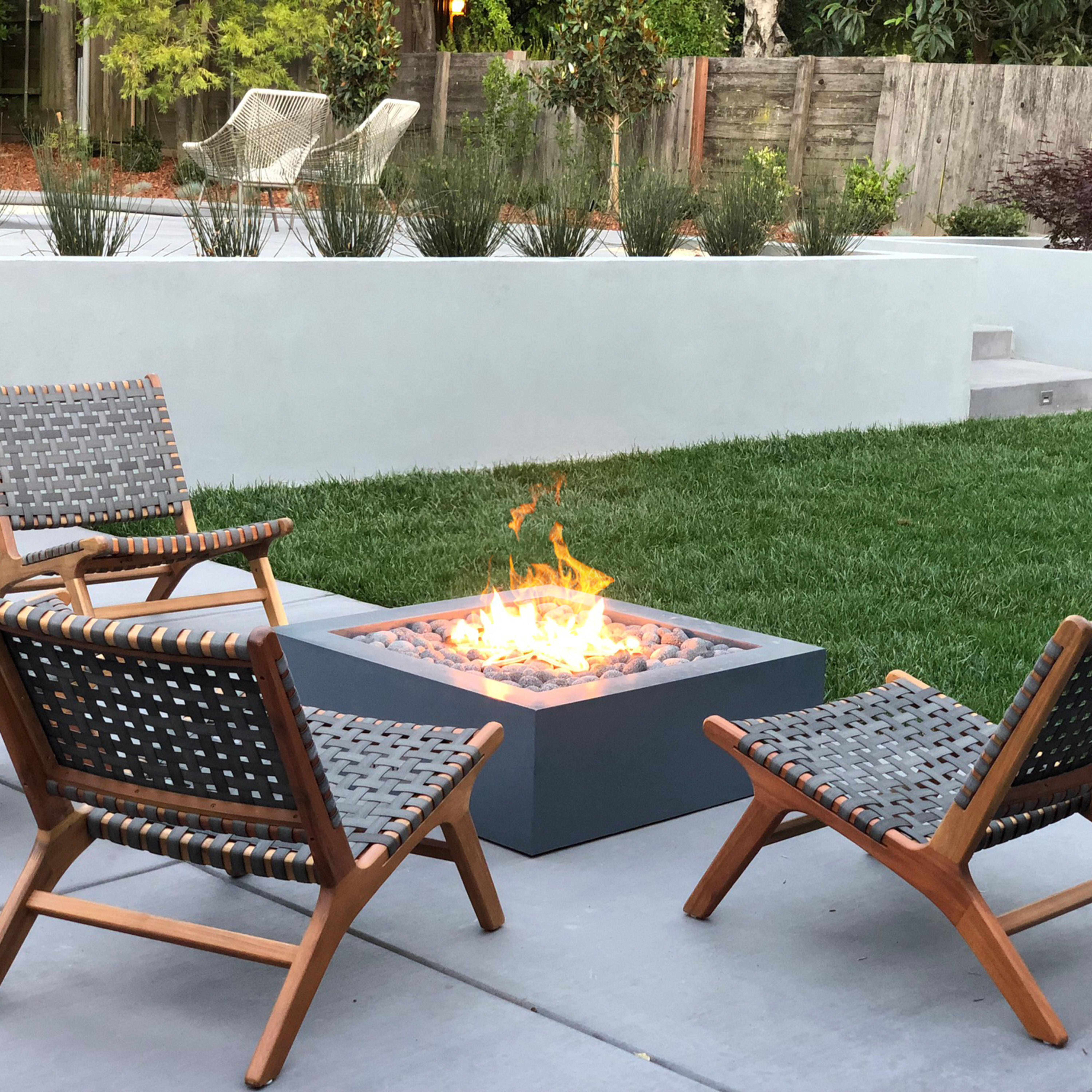 A square-shaped, concrete gas fire pit installed on a cement patio with three lounge chairs around the sides.