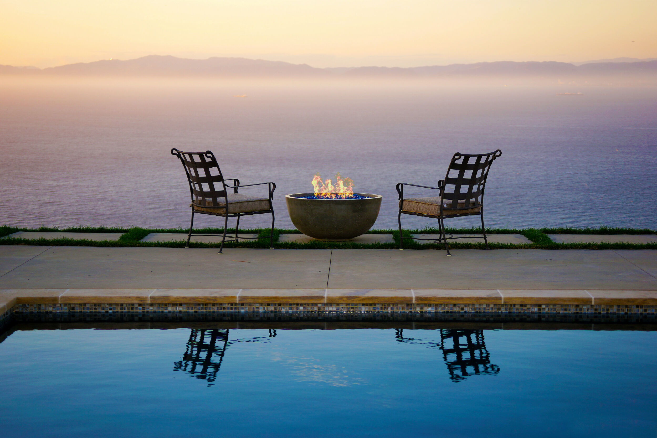 An outdoor space at dusk with two lounge chairs, a small gas fire bowl, and a lake view in the background.