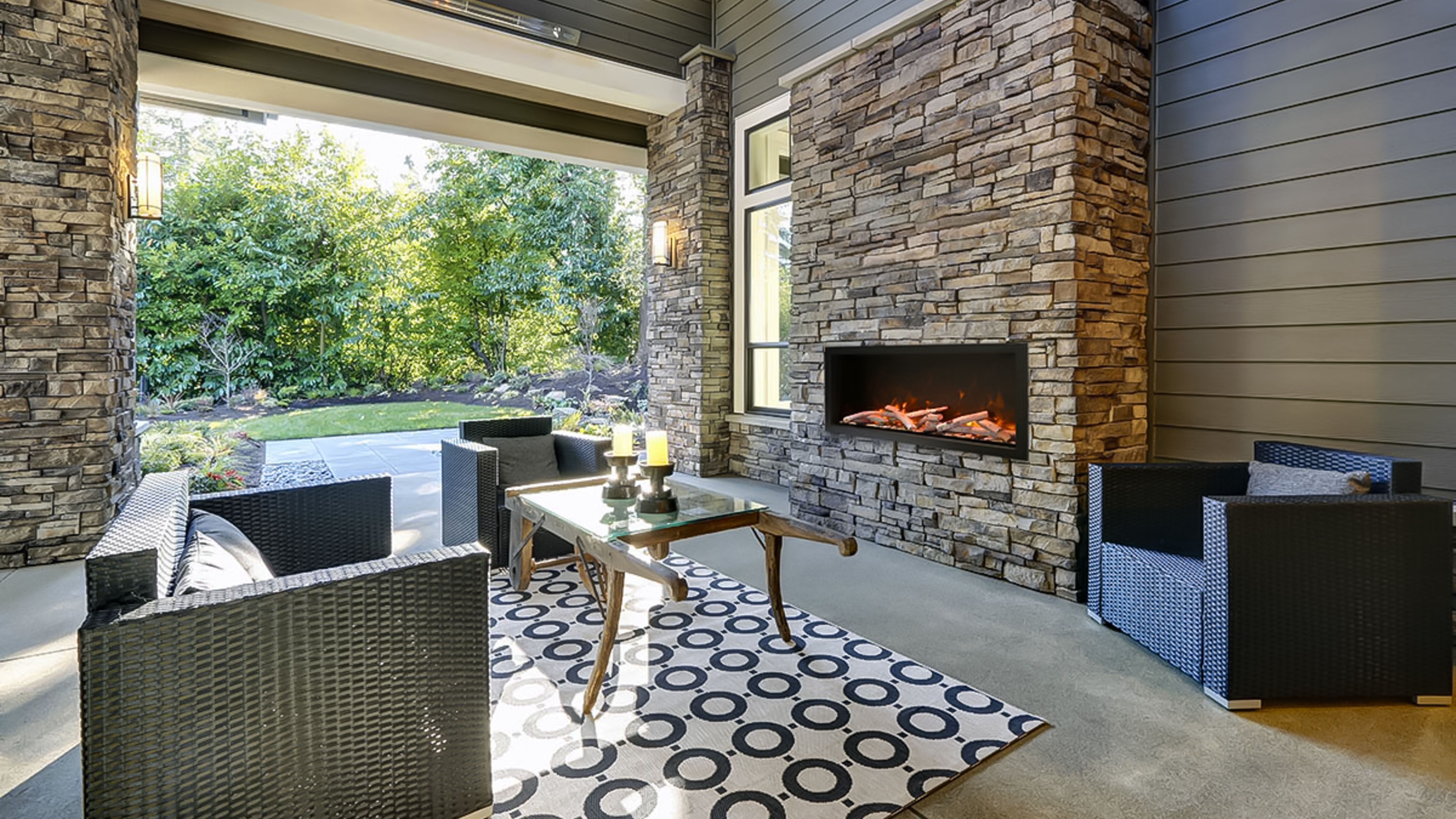 A contemporary covered patio space with a large, angular couch, a wood and glass coffee table, a white area rug with a black circle pattern, and a large natural stone hearth with a large electric fireplace and weathered, Driftwood log set.