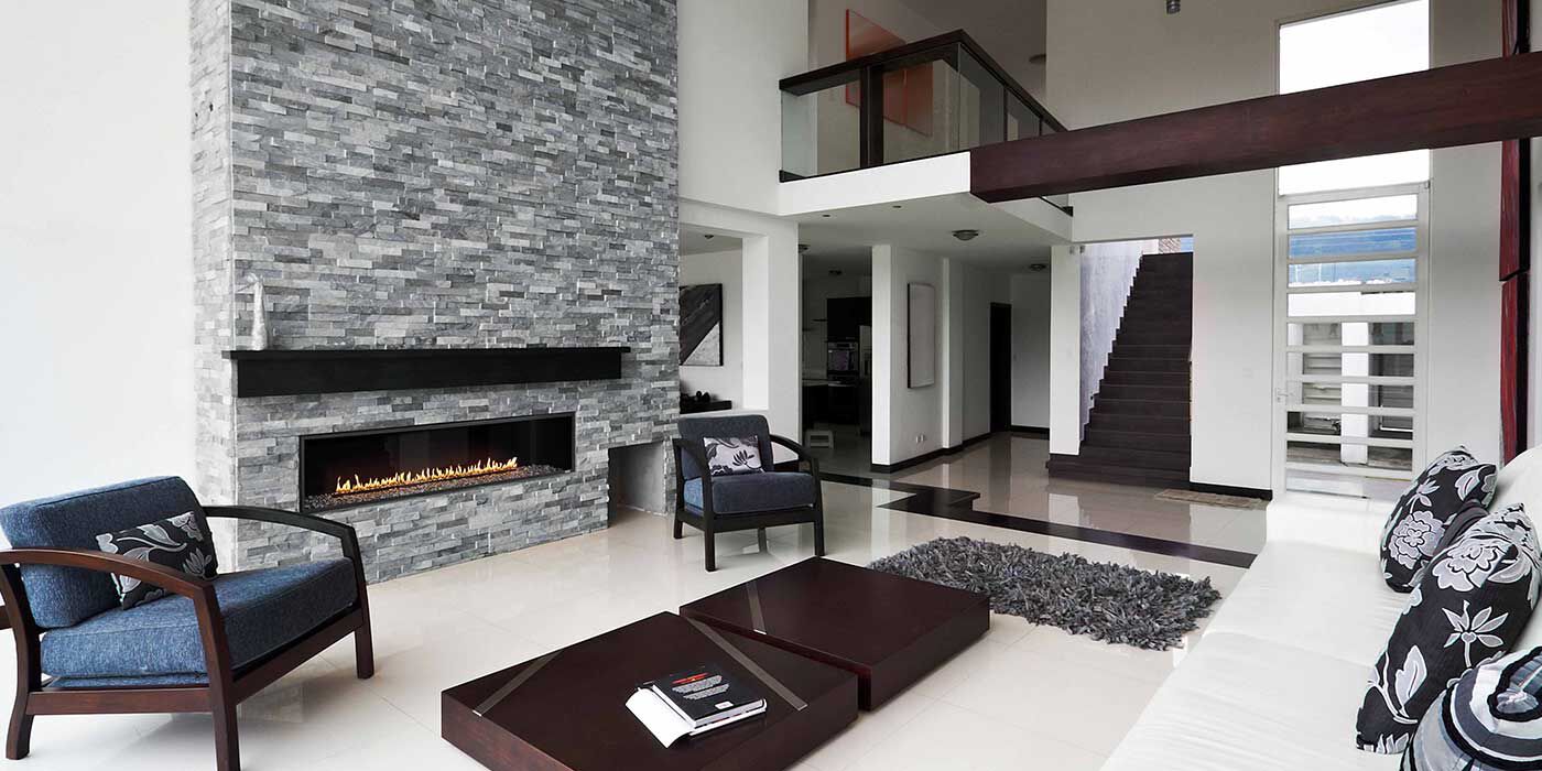 A transitional living space with an open-floor plan, white walls, dark wood trim and furniture, a navy blue accent chair, and a custom, gray stone hearth with a dark wood mantel and a large, linear gas fireplace.