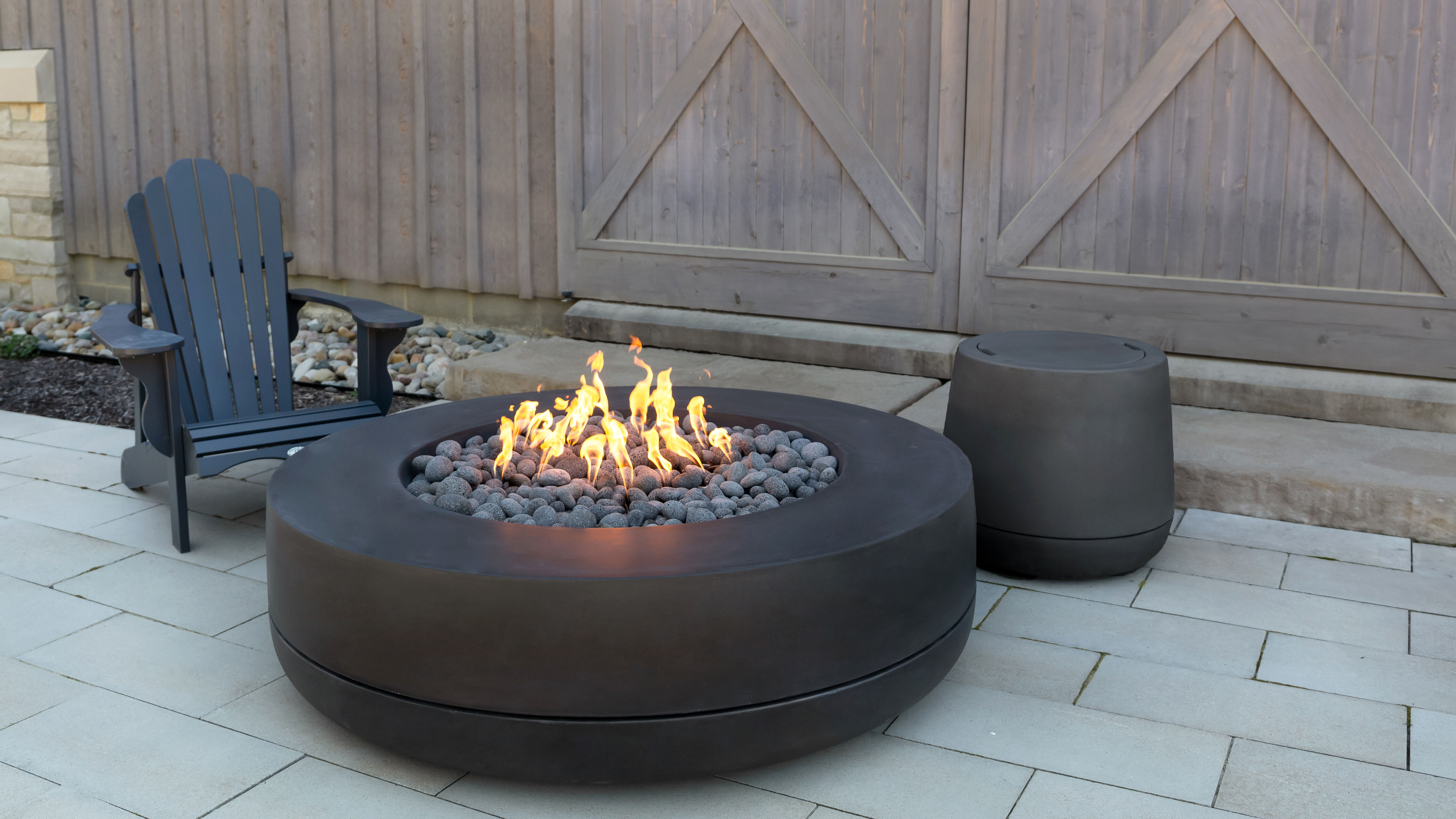The FlameCraft Tondo 60 gas fire pit installed on a stone paver patio with a matching black Propane tank enclosure and a black lawn chair. 