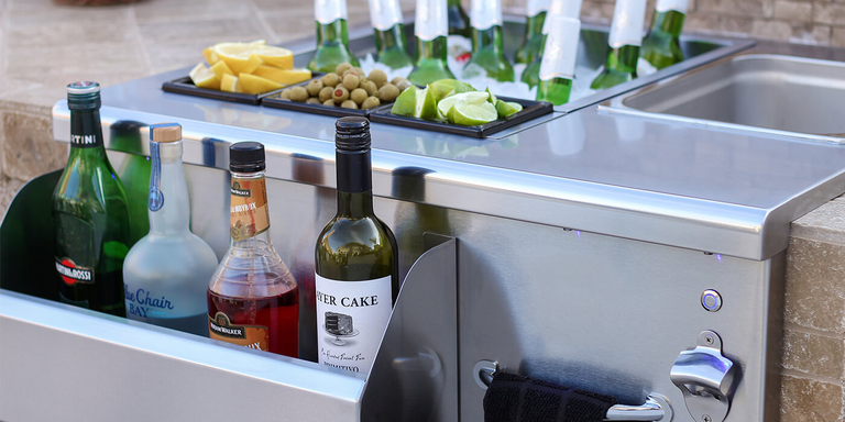 A stainless steel outdoor beverage center features a front speed rail holding bottles of wine and spirits, including Martini & Rossi and Layer Cake. The top surface includes a large ice bin chilled with bottled beers, alongside smaller garnish trays filled with lemon slices, green olives, and lime wedges. To the right, a built-in bottle opener and a towel bar with a black cloth are integrated into the unit's metal frame.