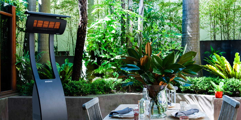A tropical looking outdoor patio space with a lot of large green plants, a dining table with chairs, and a Bromic Tungsten portable patio heater