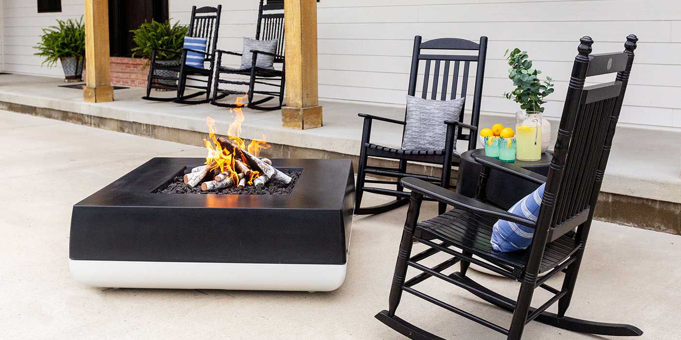 A modern farmhouse outdoor space with a wrap-around porch, black wooden rocking chairs, and a black-and-white Quadro Gas Fire Pit by FlameCraft with a matching Propane tank enclosure side table.