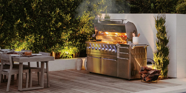 A high-end, stainless steel gas grill illuminated at night on a modern wooden deck. The grill features blue LED-lit control knobs and an open hood revealing food cooking on the grates. To the left is an outdoor dining table, and the setup is surrounded by lush green hedges and a white privacy wall.