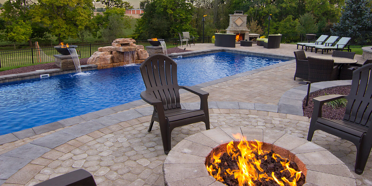 A residential backyard with a fire pit, fire and water features installed around a large in-ground pool, and an outdoor fireplace with lounge furniture around it