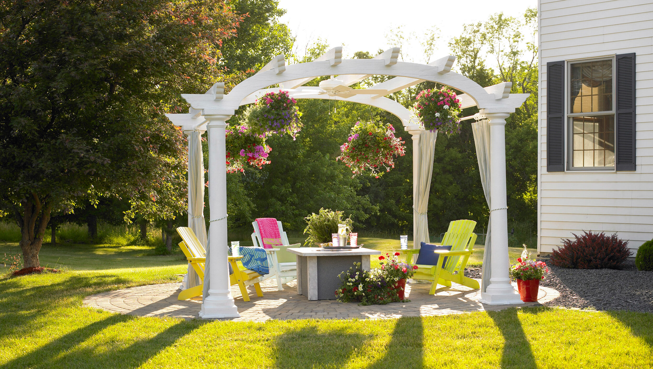 A backyard lounging space under a decorative white gazebo with three lime green plastic lounge chairs, a small gas fire table, and four baskets of flowers hanging from the rafters.