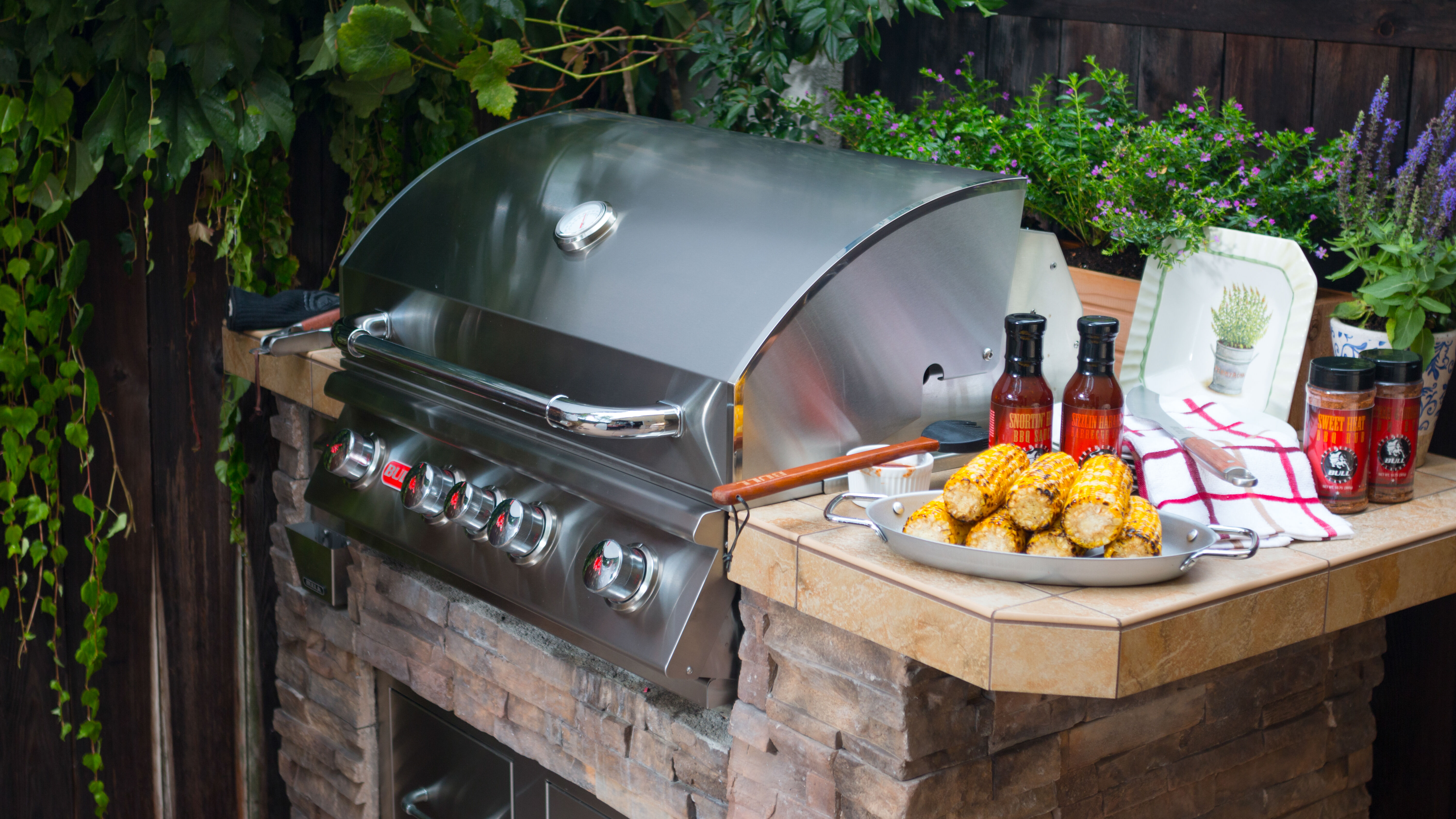 A built-in, stainless steel grill head installed on a small, natural stone kitchen island with a wooden countertop that's holding condiments, a plate of corn on the cob, and kitchen towels.