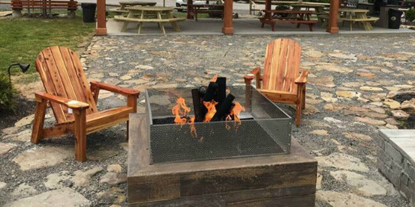 Outdoor stone patio with a square fire pit and two adirondack chairs in front of a pavillion