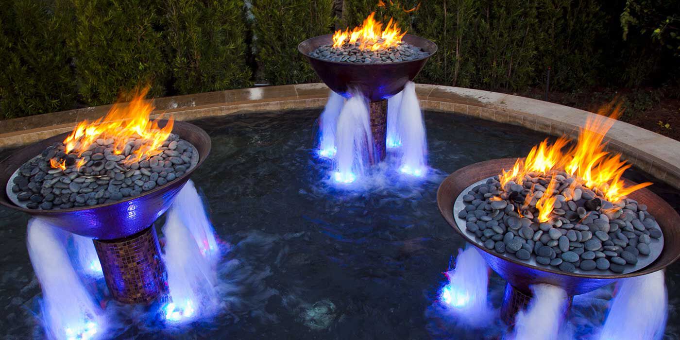 Three copper fire bowls with water pouring out of the sides installed in the center of a large, round pool.
