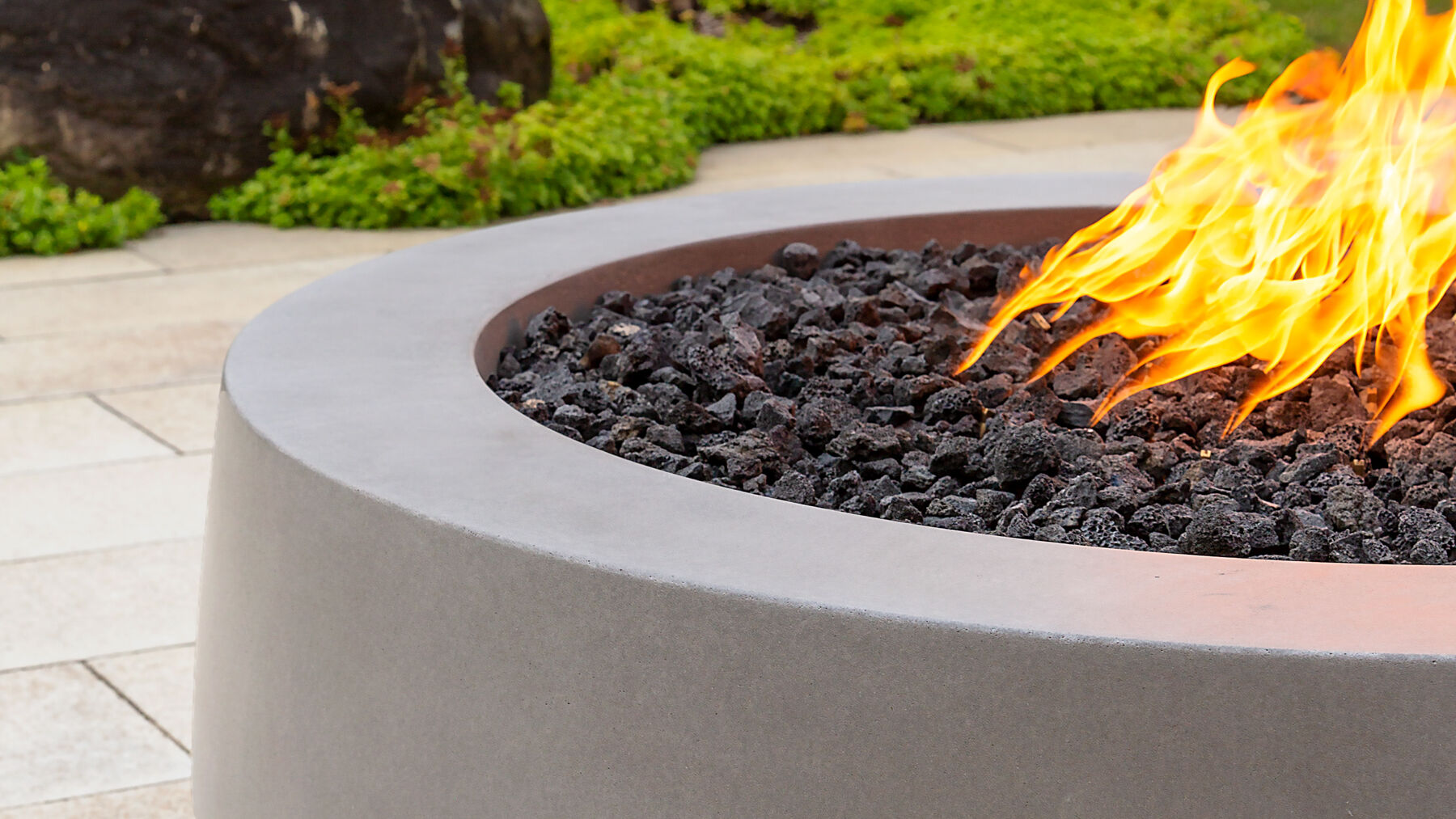 A close-up view of a gray Tondo gas fire pit from Flamecraft with black lava rock fire media.