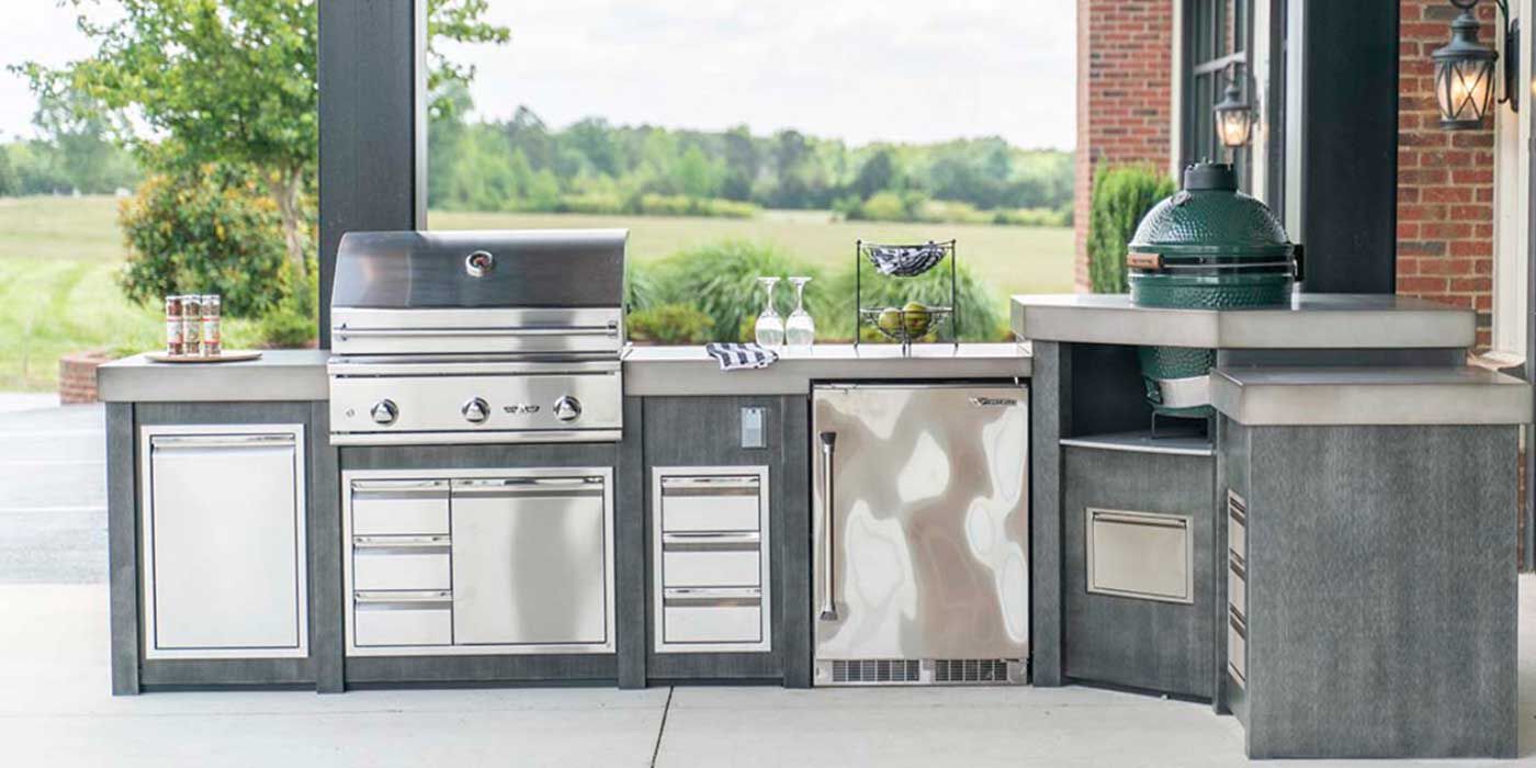 A gray and stainless steel, L-shaped outdoor kitchen island with a built-in Summerset grill and matching Summerset storage drawers, an outdoor refrigerator, and a small. countertop kamado grill.