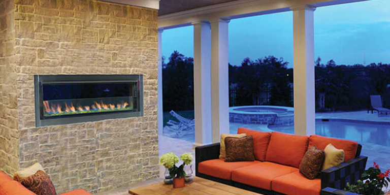 The linear outdoor gas fireplace by Superior installed on a rustic, stone wall in an enclosed patio space with orange couches, light wood furniture, and view of the background and pool in the background.