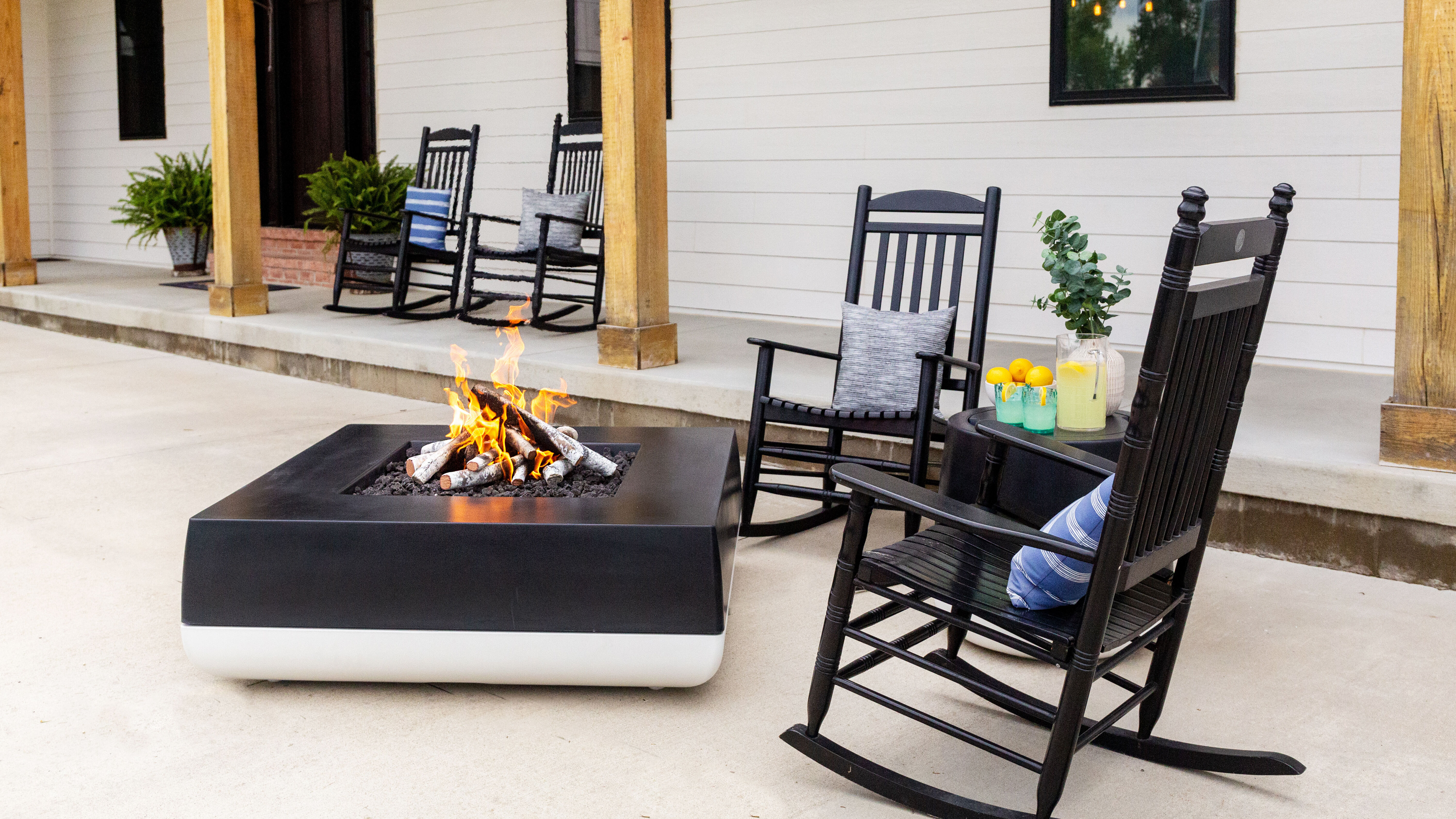 The FlameCraft Black and White Quadro gas fire pit placed on an outdoor patio with black wooden rocking chairs and a side table with refreshments.