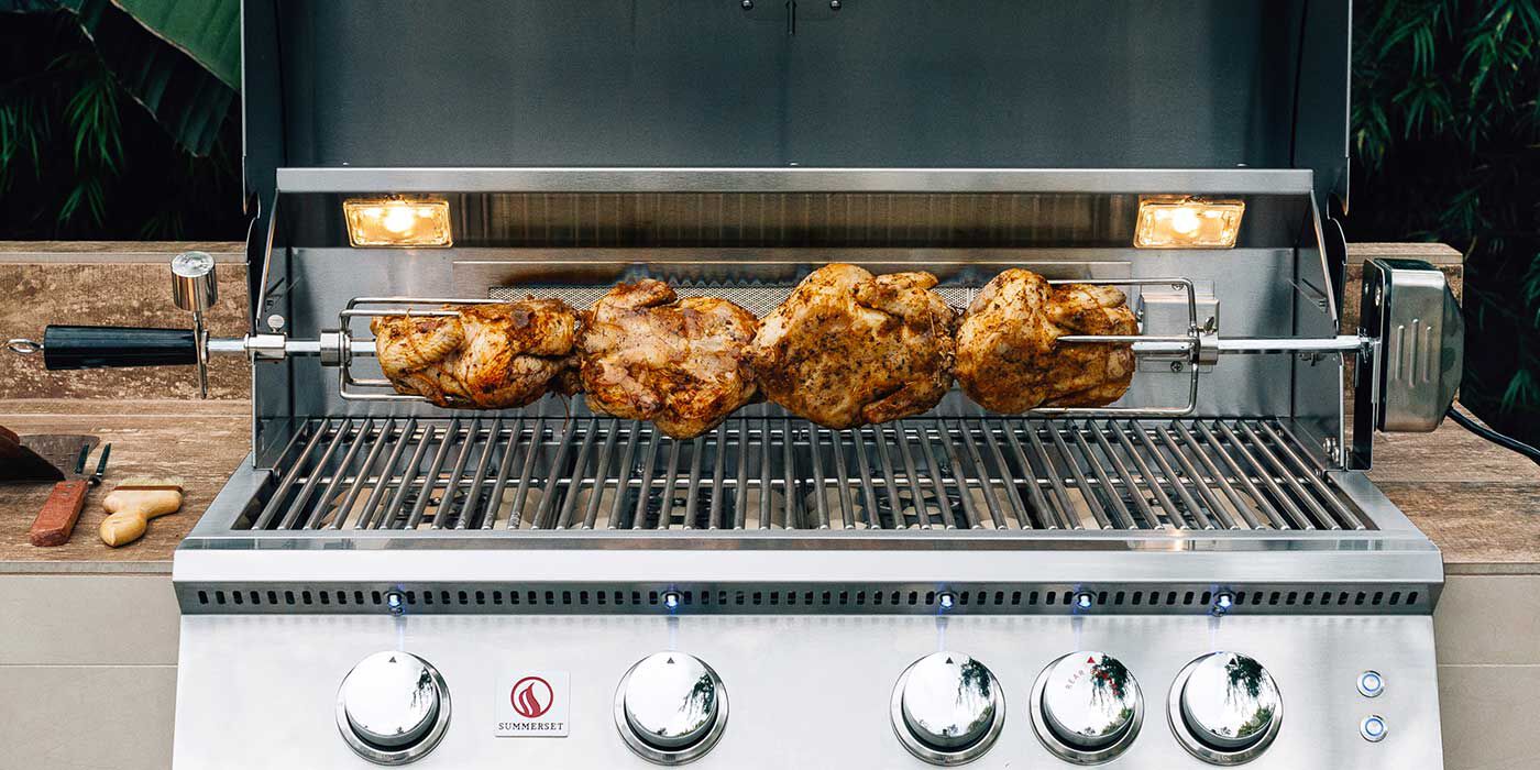 A close-up of a stainless steel Summerset grill with a built-in rotisserie spit rod four small chickens roasting over the grates.