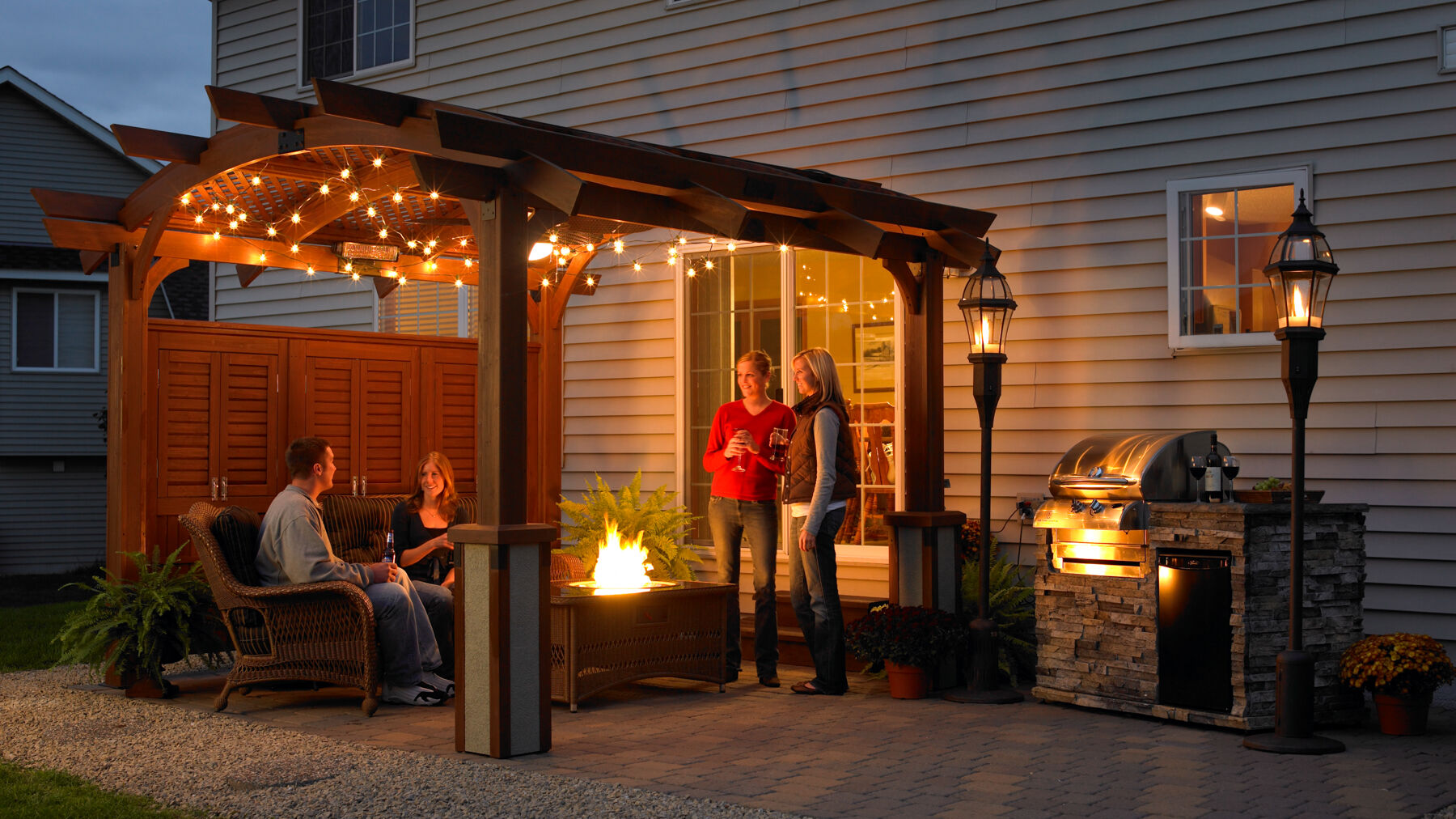 A group of people gathered on an outdoor patio at night with a wooden gazebo, a gas fire pit, an outdoor kitchen island and grill, and tiki torches.