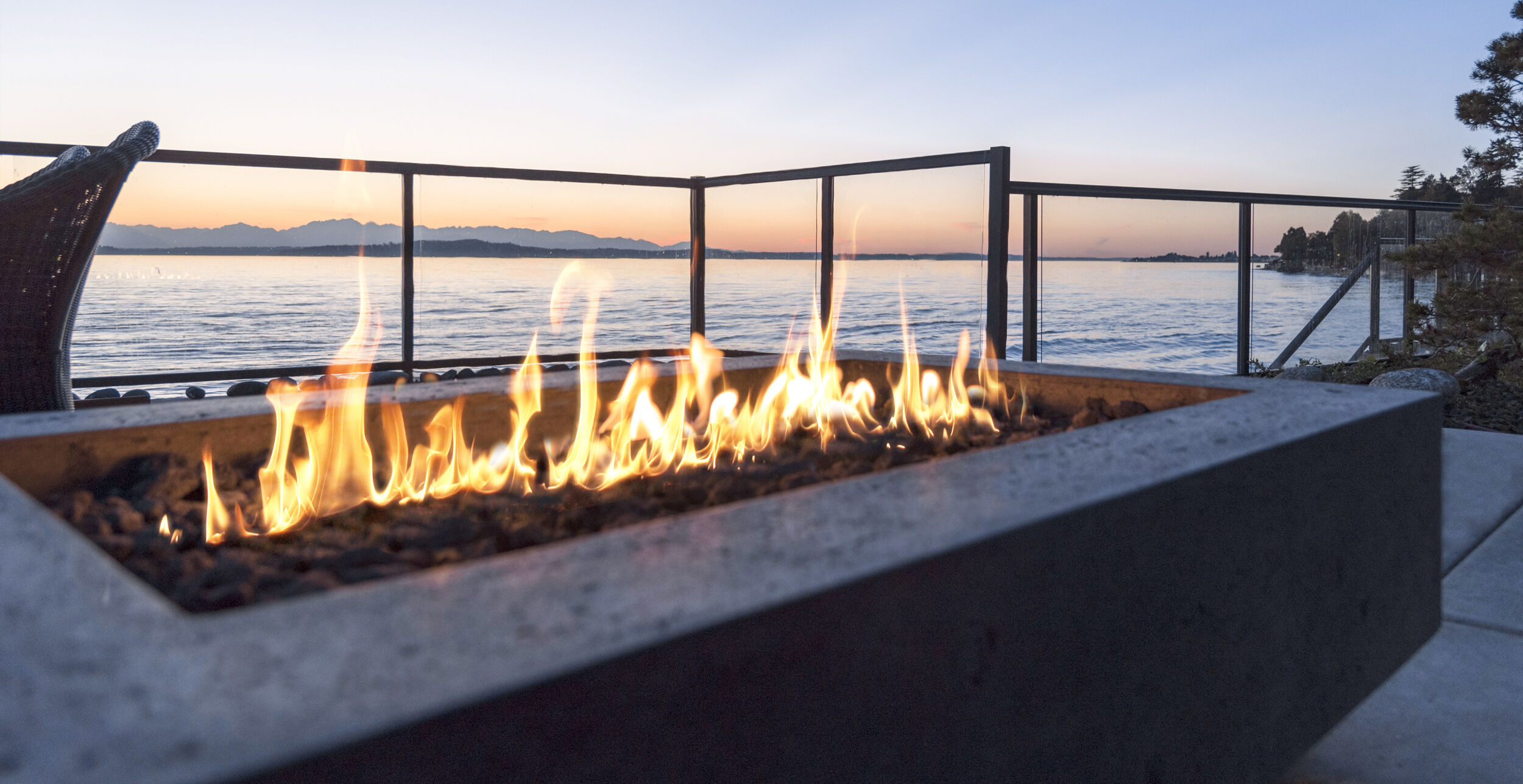 An outdoor patio at dusk with a large, linear gas fire pit and a lake view in the background.