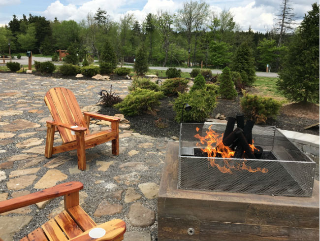 Close-up of gas fire pit and two adirondack chairs on a stone patio with a garden in the background