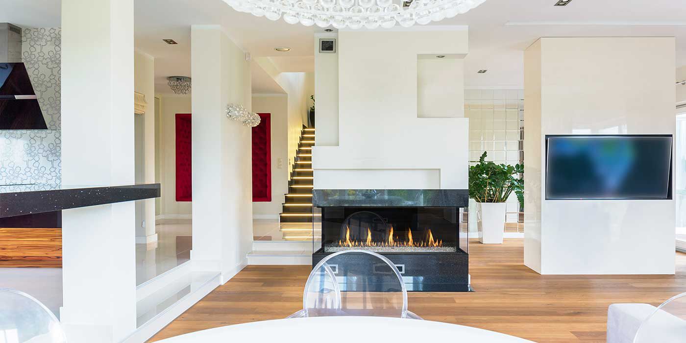 A modern living and dining room with a white, round dining table, three clear acrylic dining chairs, and wall with a flatscreen TV in the background, and a large partition in the middle of the room with a linear gas fireplace.