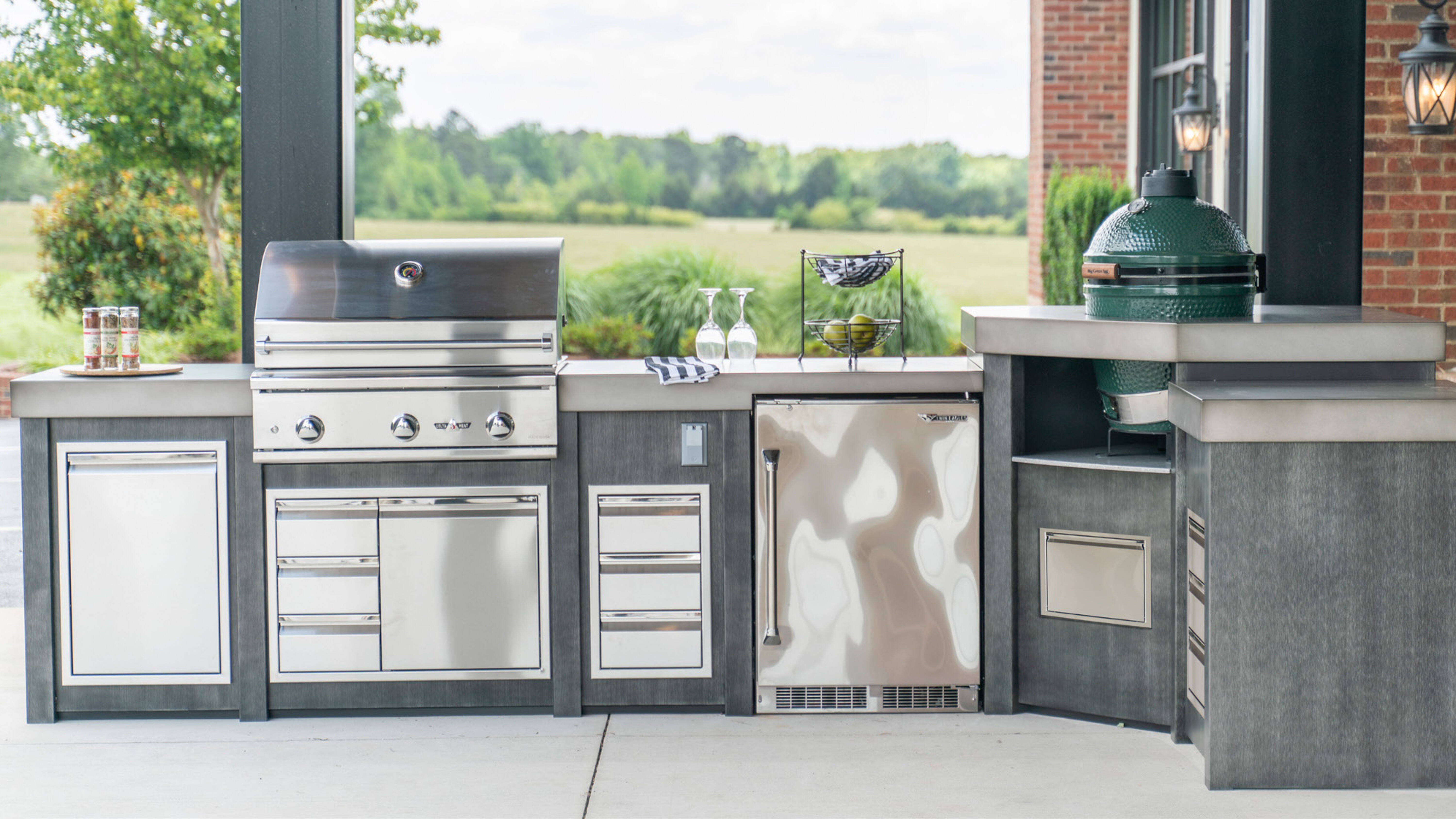 A large, curved outdoor kitchen island with a stainless steel grill head, built-in storage drawers, a refrigerator, and a kamado grill. 