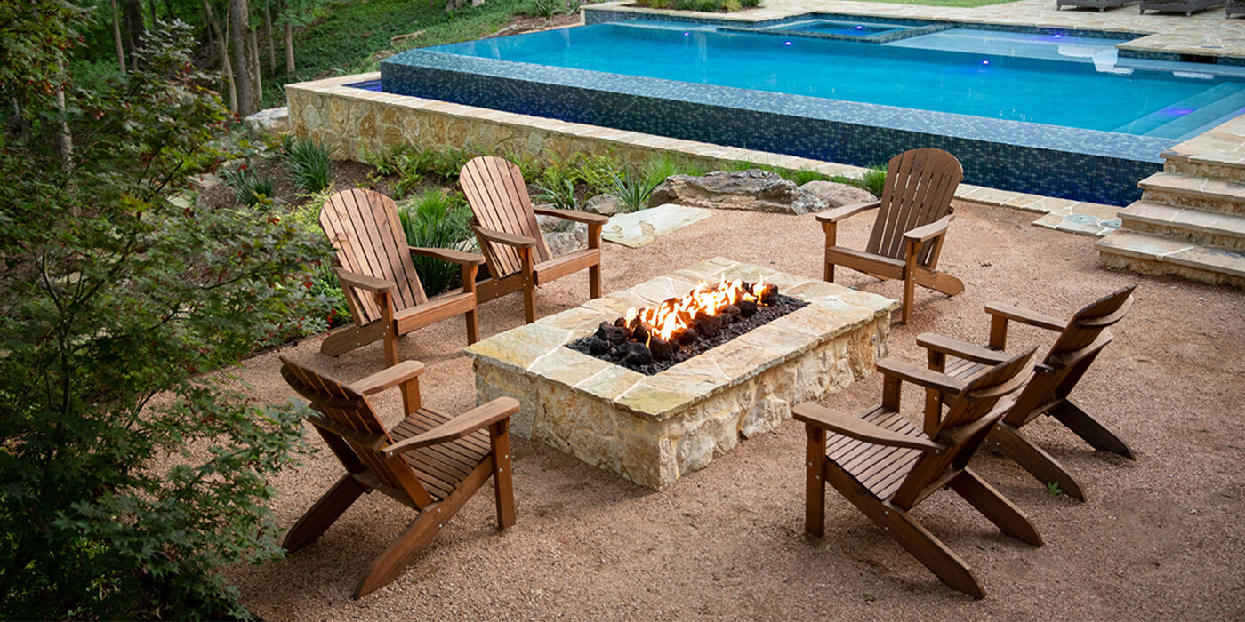 A custom-built natural gas fire pit finished with natural stone, surrounded by wooden Adirondack chairs and installed near an in-ground pool