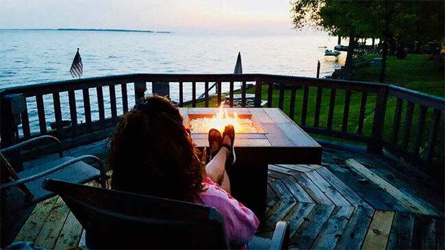 A woman sitting in front of a gas fire pit on a wooden deck with a view of the lake in the background.