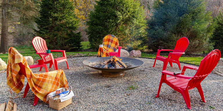 A large wood burning fire pit with four red chairs in an outdoor space surrounded by trees.
