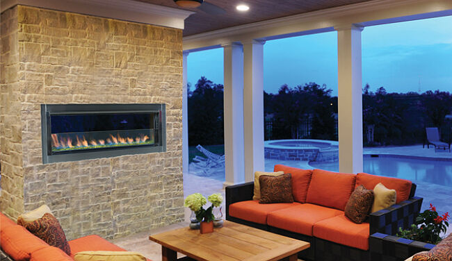 The linear outdoor gas fireplace by Superior installed on a rustic, stone wall in an enclosed patio space with orange couches, light wood furniture, and view of the background and pool in the background.