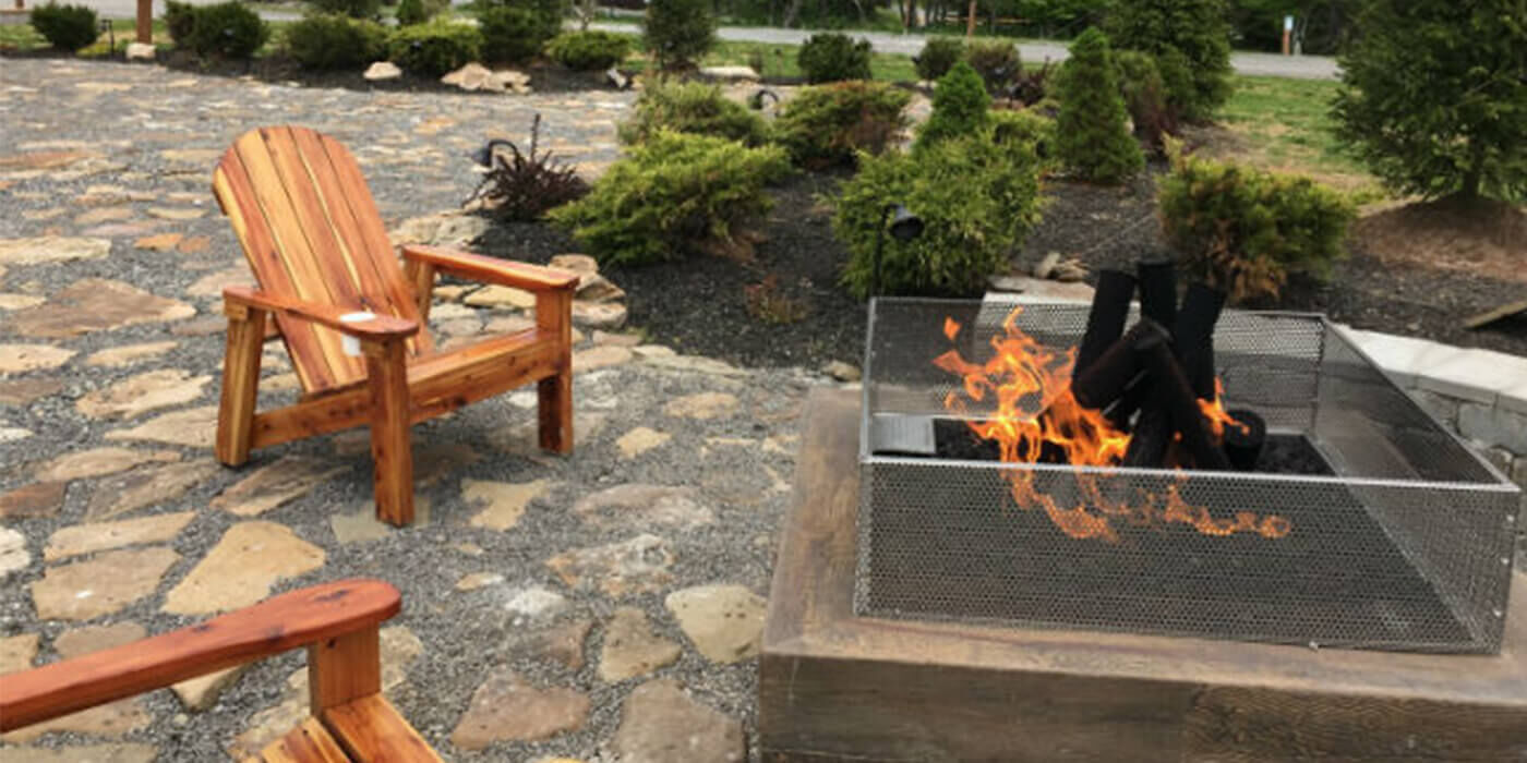 Close-up of gas fire pit and two adirondack chairs on a stone patio with a garden in the background
