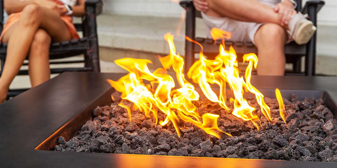A close-up view of a square, black gas fire pit with traditional lava rock media, and two people sitting down in the background.