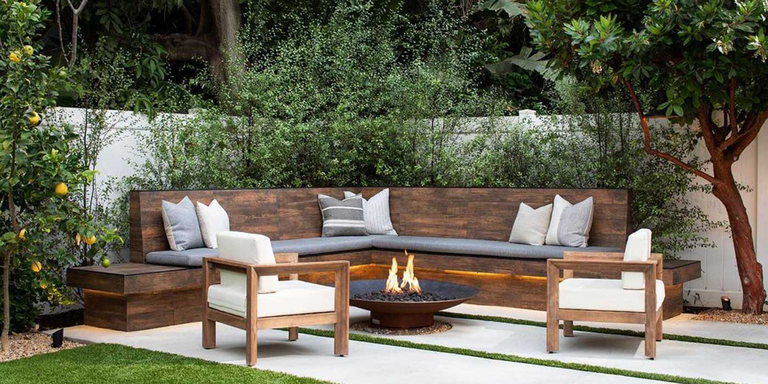 A rustic outdoor space with a wooden bench, two chairs, and a medium sized, steel gas fire pit in the center of the seating area.