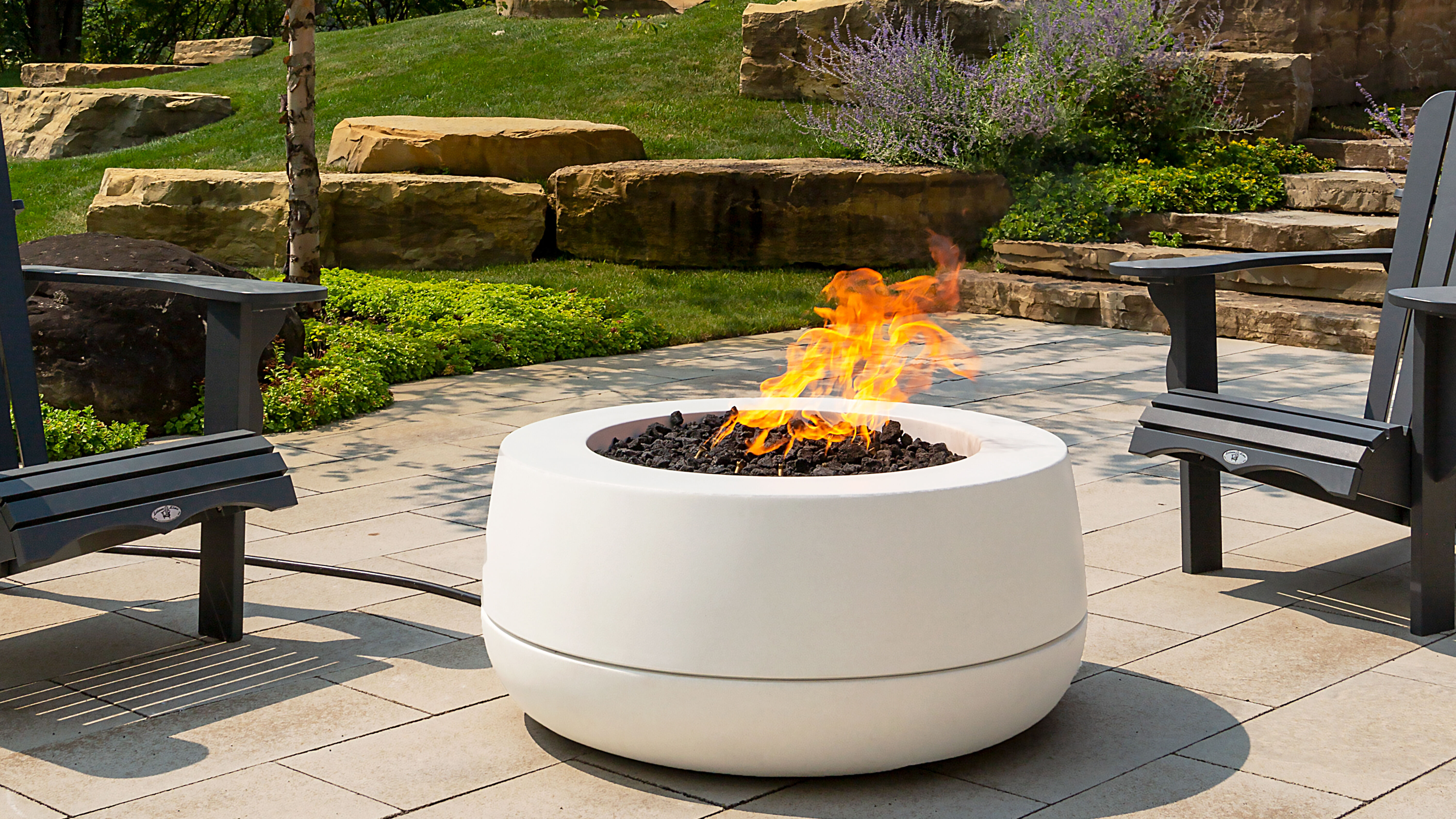 An outdoor patio space with a 30-inch Tondo gas fire pit by FlameCraft and two black plastic lounge chairs.