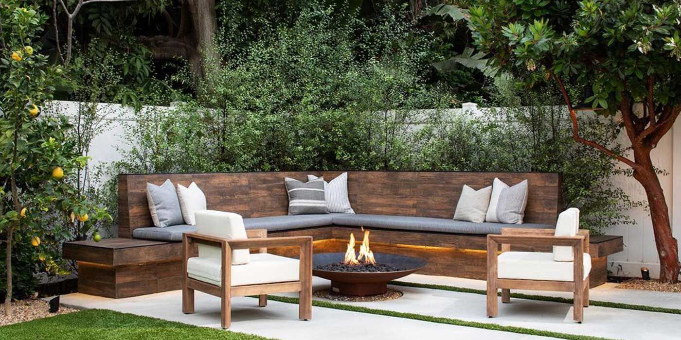 An outdoor deck surrounded by lush green foliage with a large, L-shaped blue couch and a black gas fire table.