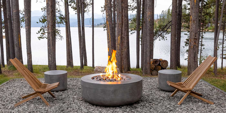 A Prism Hardscapes Curva gas fire pit installed on gravel-paved platform in a wooded outdoor area