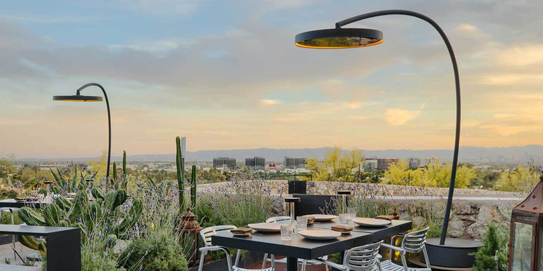 A high-end outdoor dining space on a dock with three tables, black chairs, green ferns in the background, and two portable gas patio heaters positioned over the tables.