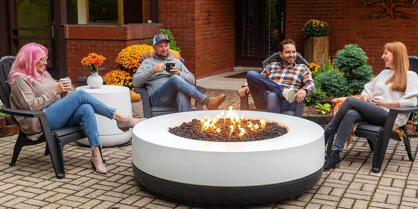 Two women and two men sitting in plastic lounge chairs around a large, round, black and white Tondo 60 gas fire pit.