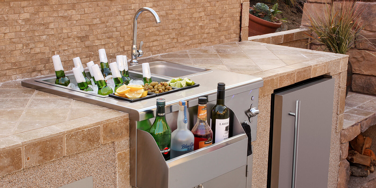 An outdoor kitchen bar setup featuring a built-in stainless steel sink and a larger well filled with ice and bottled beverages. Small trays of lemon wedges, olives, and lime wedges are set between the two wells. Below, a front-mounted metal rack holds several liquor and wine bottles. The bar is integrated into a stone tile and stucco structure, with an integrated stainless steel door and fireplace-like features to the sides.