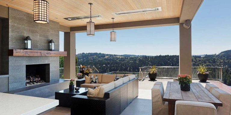 An outdoor living space with a seating area in front of a fireplace, a dining area with chairs, hanging light fixtures, and two Bromic Platinum heaters recessed into the ceiling