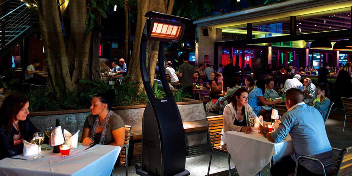 An outdoor restaurant patio at night with a large, freestanding Bromic Tungsten patio heater in the center