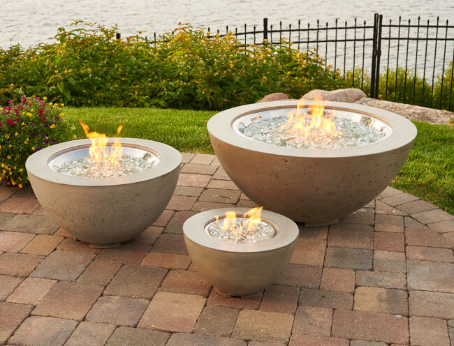 A small, medium, and large sized Cove Gas Fire Bowl on a brick patio with green bushes, a black fence, and a view of the lake in the background.