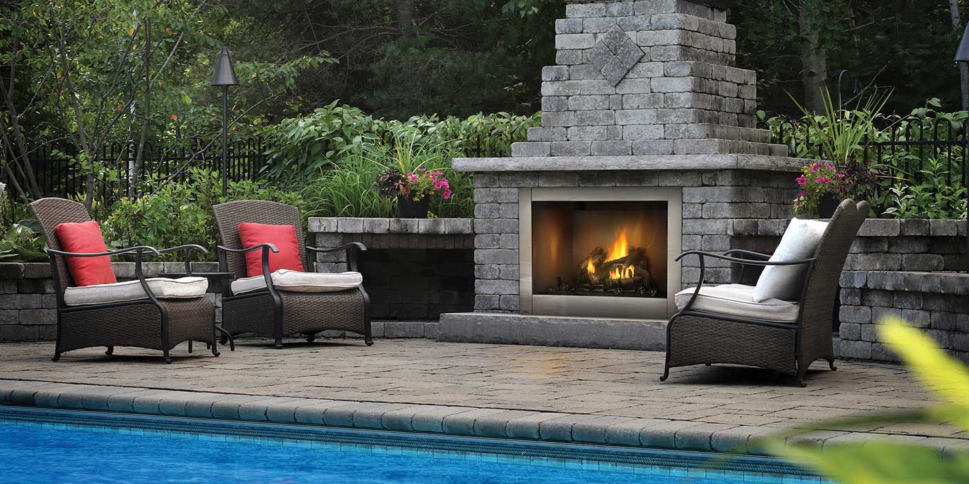 An outdoor lounging area by a pool with two chairs, a couch, and a traditional masonry outdoor fireplace.