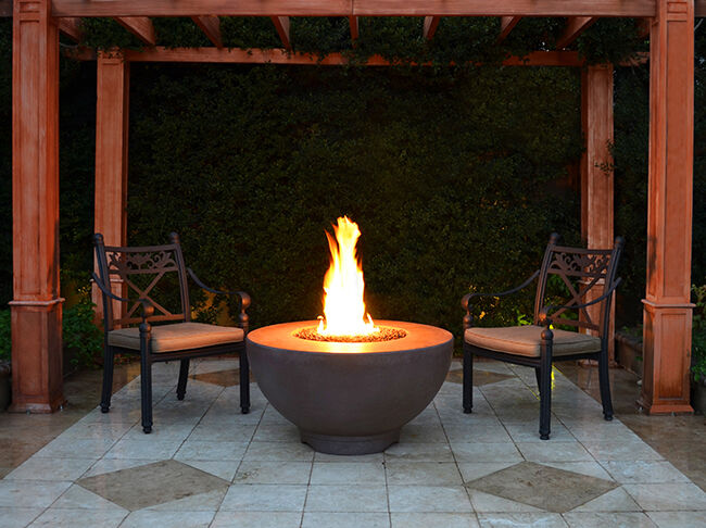 The Sienna Gas Fire Pit installed on a stone patio with elegant, wooden pillars and two chairs.