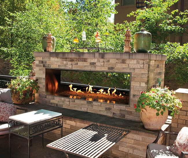 The Carol Rose Linear See-Through Fireplace by Empire install outside on an open patio, finished with neutral-colored bricks and surrounded by green shrubs, trees, plants, and potted flowers.