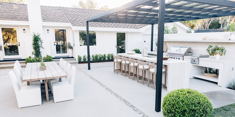 A wide-angle view of a luxurious, all-white outdoor living space. A long minimalist island with a sink and barstools sits under a black pergola, while a separate dining table with white chairs occupies the concrete patio nearby.