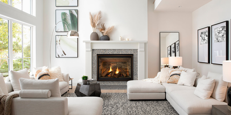 Modern living room with a cozy fireplace, surrounded by white sofas and cushions. Large windows let in natural light, and framed art decorates the walls.