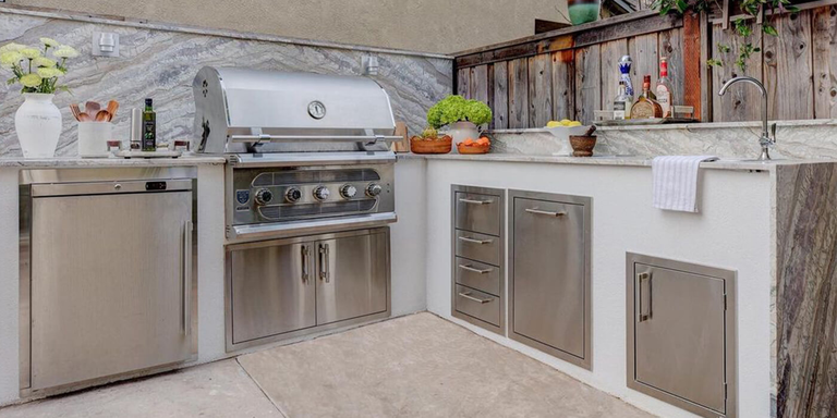 A compact L-shaped outdoor kitchen featuring white cabinetry and stainless steel storage drawers. The setup includes a large professional grill and a sink, set against a decorative gray marbled backsplash and a wooden fence.