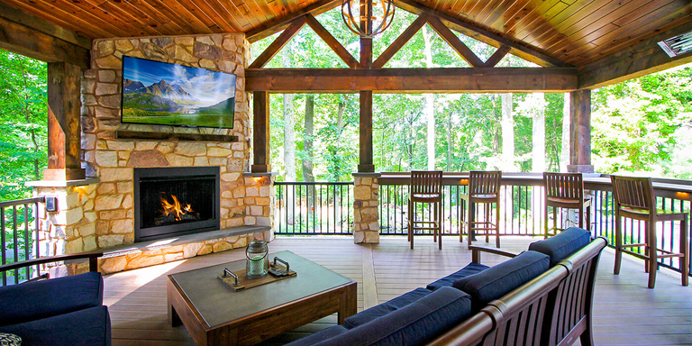 A rustic covered porch features a large stone fireplace with a TV mounted above it, a comfortable dark blue sofa, and a coffee table. Large dark wood beams and a vaulted plank ceiling enclose the space, which overlooks a lush green forest. Along a deck railing, four bar stools are lined up at a counter, perfect for dining with a view.