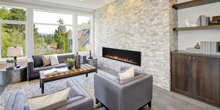 Modern living room with large windows, a stone accent wall, sleek fireplace, and cozy gray furniture. Bright, inviting atmosphere with a neutral palette.